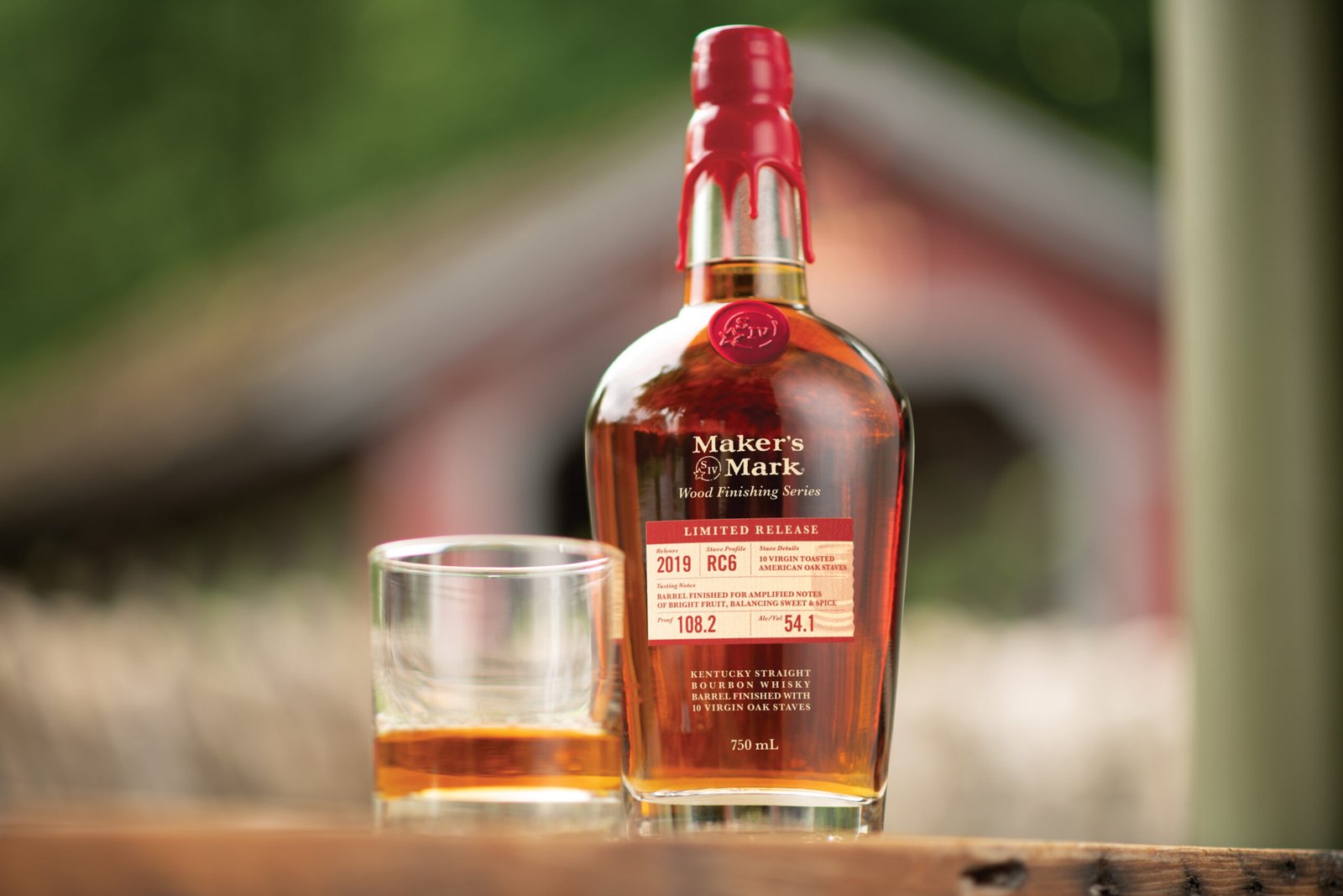 A whiskey bottle and glass photographed in front of a red Kentucky barn