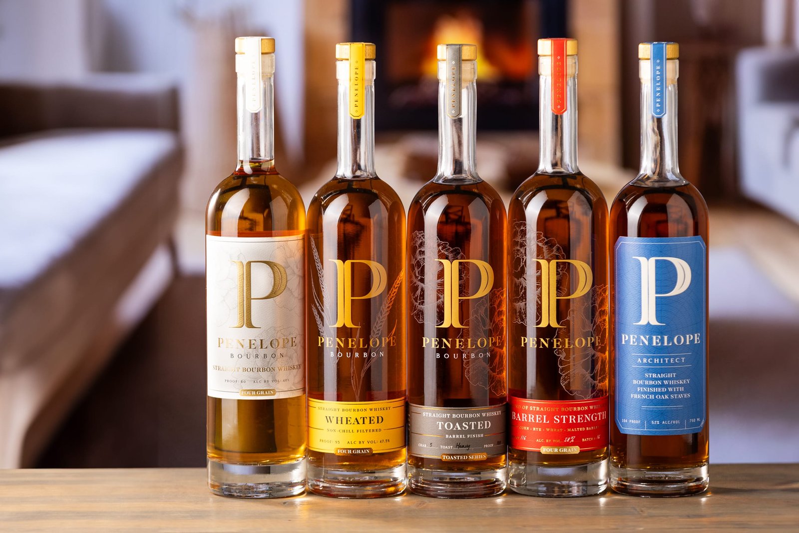Five Penelope bourbon bottles lined up on a wooden table in front of a warm open fire