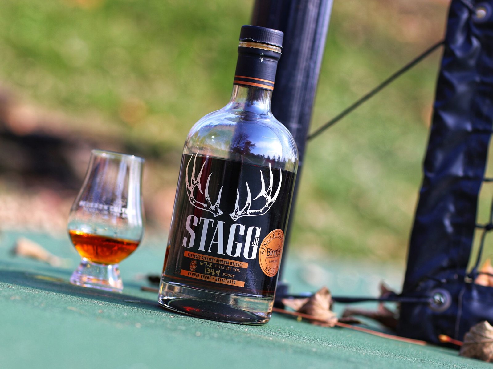 A bottle of Stagg Jr single barrel bourbon beside a filled glass on a wooden rail outdoors